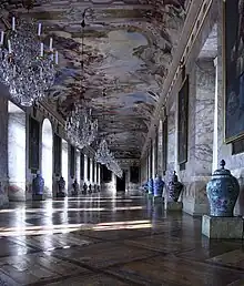 A view down the Ahnengalerie. The walls, made of scagliola, are lined with portraits of the rulers of Württemberg, their arms, and an urn. Above the gallery is a massive fresco, featuring several Classical figures, glorifying the reign of Eberhard Louis.