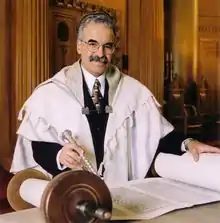 A gray-haired man with a full mustache faces the viewer, wearing a large skullcap, round wire-rimmed glasses, black suit with white shirt and patterned tie, and a full white prayer shawl with light gray stripes. On a table before him is an open Torah scroll; his left hand rests on one side of the scroll, and his right hand holds a silver pointer pointed at the words written on the scroll. A wall and part of a closed Torah ark are visible behind him.