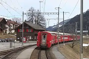 Two-story wooden building with gabled roof next to double-track railway line with red train on one track