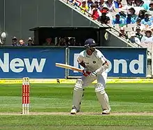 An Indian cricketer in a blue helmet, waiting to receive a delivery. He is standing in front of his wicket, with spectators in a stand behind him.