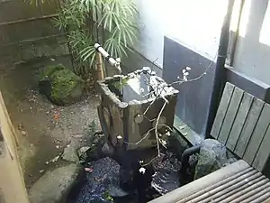 Square concrete basin, tall and slightly flared, decorated with circles; an ABS plastic tap drips into it. Bamboo, a bare branch laid on the sink, and a sump pond with a black plastic downspout from the roof.Takesunoko-en (竹簀の子縁) for drainage (see engawa)