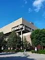 A person biking past the corner of a brick and concrete building, under a nearly clear sky. In front of the building are several trees and bushes, which themselves are behind a large section of pavement.