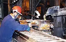 A man in a hard hat with a clear face plate leans over a conveyor belt with a metal instrument. He is in a factory setting with heavy machinery in the background.