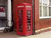 A red, cast iron telephone kiosk with a domed roof. it stands against a red brick wall at the corner of a building.