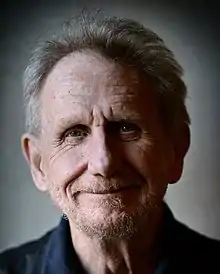 A silver-haired man with receding hairline and short beard and mustache smiles at the camera