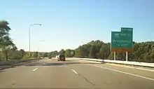 Ground-level view of three lanes of a divided freeway; a large, green exit signs is visible to the right, and trees surround the freeway on both sides.