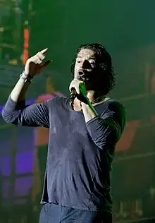 A man with blue jeans and shirts half-opened holding a microphone with his right hand while singing in front of a stage, with a city simulated background behind him.