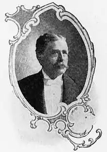 Head and shoulders of a white man with mustache wearing a tuxedo and white bow tie. The portrait is surrounded by a decorative oval-shaped frame.