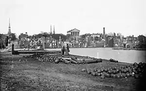 A single soldier stands among cannons and cannonballs across a river from the ruins of a city.