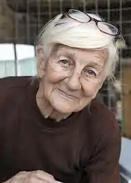 Photograph of an elderly, grey haired woman with glasses perched on top of her head, leaning to her left and wearing a brown sweater.