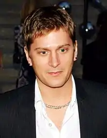 A young man with short brown hair wearing a dark jacket and a white open-necked shirt