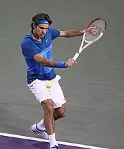 A brown-haired male tennis player with white shorts, a blue shirt and a blue headband swings a right-handed forehand on a hard court surface