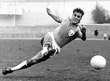 A black-and-white photograph of an association football player caught dramatically in mid-motion. As he kicks the ball with his right foot, his face fixed in concentration, he is about to fall to the ground on his left side.