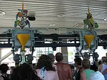 The Roller Soaker's station is seen with overhanging Hershey character's refilling the ride vehicle's water. Guests waiting in the queue line observe the roller coaster.