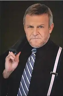 A man with dark/grey hair, wearing a black shirt with pink/blue tie.