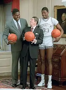 Two tall African-American men, one in a suit, one in a gray basketball uniform, stand behind a shorter elderly white male in an ornate room, with each man holding a basketball.