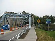 A blue metal truss bridge crosses over a small creek bordered on each side by sloping retaining walls made of stone.