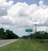 A color photograph of an empty two-lane highway disappearing into the distance, lined by trees on both sides and a field to the right; at the center is a green sign that reads "Rosewood"