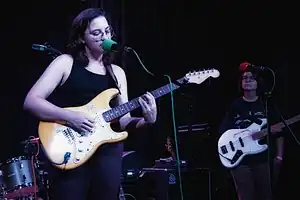 Tucker in a black tank top playing guitar and singing on a stage; a bandmate is behind and to the right