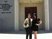 Man with light skin and dark hair and woman with dark hair and light skin stand in front of a building with a sign that reads "The Church of Jesus Christ of Latter-day Saints; Visitors Welcome"