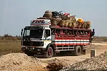 A flatbed truck with a roof over its bed drives down a dirt road. Beneath the roof sit quite a few people on beches; two hang on to the back of the truck. Atop the roof is a variety of goods in woven baskets. It says "Transport" on the front of the truck.