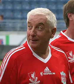 The head and upper torso of a gentleman in his late 50s. He has short white hair and is wearing a red football shirt, which has the Liverpool F.C. crest on the left breast, and a crest on the right breast that says "Liverpool Legends". A white logo of the Adidas sponsor is visible in the centre of the shirt, and three white stripes are present on the shoulder.