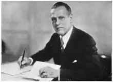 Black and white photograph of a man wearing suit holding a pen with one hand and a piece of paper with the other.