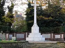 A white cross standing on a plinth and steps and behind is a wall with inscribed plaques