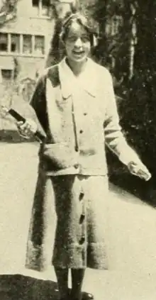 A smiling young white woman photographed outdoors, wearing a cardigan suit over a white blouse with a long pointed collar; she holds books with one arm