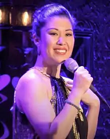 Miles in an off-the shoulder concert dress, smiling at the camera and holding a microphone