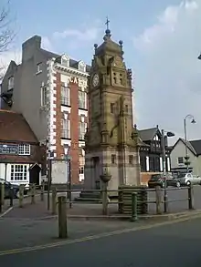 A square structure in banded stone with various decorations, including a statue on a column.  On one face is a clock and at the top is a short spire with ball finials.