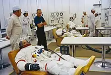 Several men in space suits recline on couches as others stand by.