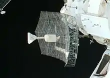 A rectangular dish shape of scaffolding covered in transparent sheeting, with a white insulation-covered radio receiver and support projecting from the centre. The blackness of space serves as the backdrop.