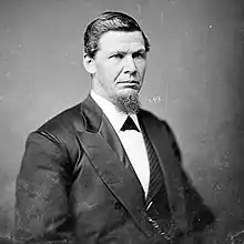 A stern-faced man in a dark suit looks straight ahead. He sports facial hair on his chin.