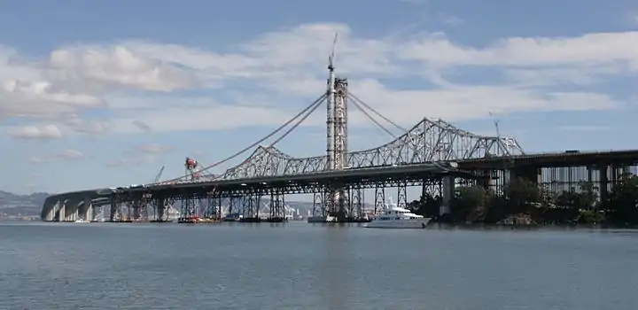 October 1, 2011: In the distance, the Left Coast Lifter is placing the last of the main span deck segments. Two additional short segments will join the main span to the curved skyway extension. The main suspension cables will follow the curves outlined by the recently installed catwalks. Ten holdback cables to the right of the tower (below the catwalks) preload the tower, bending it 17 inches (430 mm) west against the forces to be imposed by the main cable when the bridge is complete, allowing the tower to be vertical when the holdbacks are removed. Subsequent to this image the traveler support cabling and cabling supports were installed, and all of the main cable strands have been placed and compacted, the suspender cables hung, attached, and tensioned.