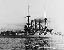 A large gray warship sits in calm water, with dozens of men standing on its main deck.