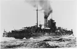 A large battleship steaming away through the water at high speed. Its stern can be seen in the foreground with water hitting the sides of the ship. Smoke can be seen billowing out of the funnels of the ship.