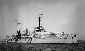 A large light gray warship sits motionless in harbor. Two tall masts stand in the center of the ship.