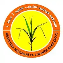 Round logo of a grass plant against a yellow background, with the organization's name in Kurdish and Arabic on an orange border