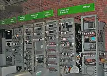 A series of seven tall metal racks filled with electronic equipment standing in front of a brick wall. Signs above each rack describe the functions carried out by the electronics they contain. Three visitors read from information stands to the left of the image