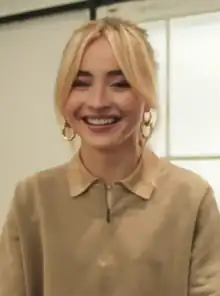 A young woman with bleached hair, large double-hooped earrings, and dark eyebrows smiles.