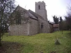 A flint church seen from the northeast showing the chancel, nave and aisle with a catslide roof, porch and battlemented tower