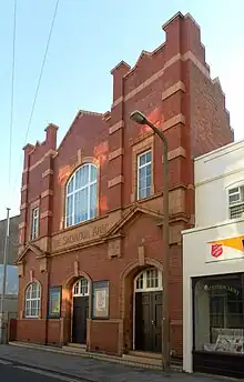 A tall building with deep red brickwork and yellowish stone dressings. There are two identical entrances: wooden doors in recessed stone doorways with prominent yellow keystones.  A window to their left is in a similar recess.  Two pediments flank a stone frieze with the words "THE SALVATION ARMY".  The upper floor has two rectangular windows and a central arched window with another keystone.