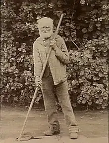 old bearded man wearing trousers and a coat, leaning on a pole