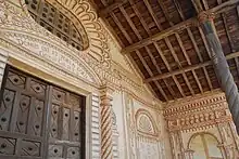 A wooden door of the main entrance to the church and wooden roof of the front porch. The whitish wall is decorated with floral motives painted in orange and a large oval window surrounded by flower petals is located above the door.