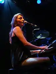 A woman with long dark hair playing a piano and singing into a microphone