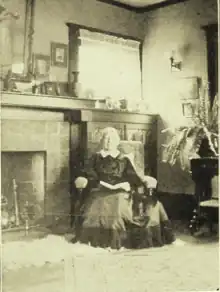 An older white woman, seated indoors, next to a fireplace, with a book open in her lap; she is wearing a long dark skirt and has white hair