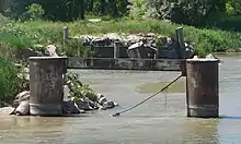 Two vertical cylindrical pilings, joined by horizontal beam near tops, standing in river