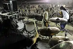 Two male guitarists and a drummer performing on stage in front of a small crowd