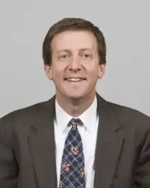 A portrait shot of Scott S. Harris, looking straight ahead. He has short brown hair and is wearing a charcoal blazer with a blue patterned tie over a white collared shirt.